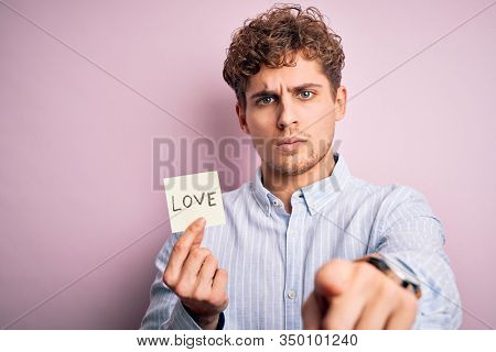 Young blond romantic man with curly hair holdiing paper with love message for valentines day pointing with finger to the camera and to you, hand sign, positive and confident gesture from the front