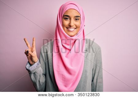 Young beautiful brunette businesswoman wearing pink muslim hijab and business jacket smiling with happy face winking at the camera doing victory sign. Number two.