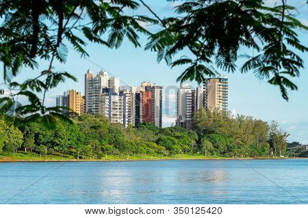 View Of A City Lake Through The Tree Leaves. Lake Surrounded By Nature And Few Buildings On The Back