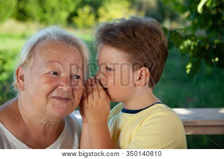 Boy whispering to grandma in summer outdoors garden, grandkid and grandmother lalking.