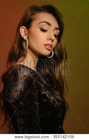 Attractive Brunette Woman In Stylish Jewelry And Black Velvet Dress, Posing Sideways On Colorful Bac
