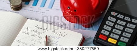 Close-up Of Red Piggy Bank Terminal And Notebook With Notations. Monthly Expenses On Rent Car Food A