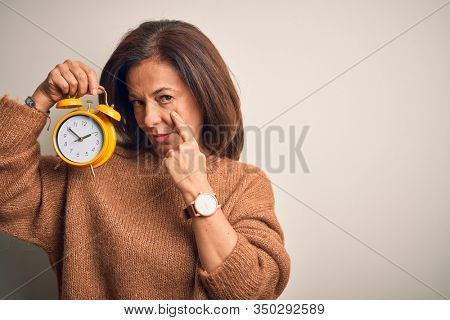 Middle age brunette woman holding clasic alarm clock over isolated background Pointing to the eye watching you gesture, suspicious expression