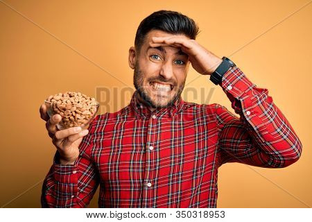 Young handsome man holding bowl with healthy peanuts over isolated yellow background stressed with hand on head, shocked with shame and surprise face, angry and frustrated. Fear and upset for mistake.