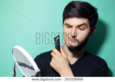 Studio Portrait Of Young Man With Half Shaved Face, Looking In Mirror Holding Electric Shaver Trimme