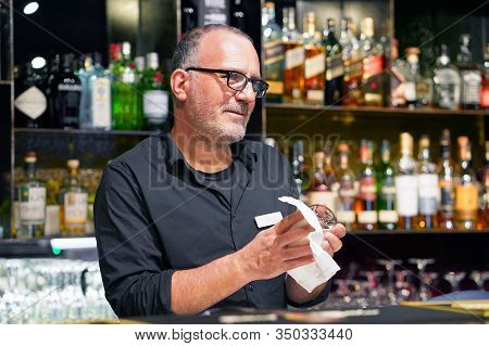 Elderly Bartender Cleans The Glass. A Handsome Bartender Polishes A Glass Of Wine Glasses. The Conce