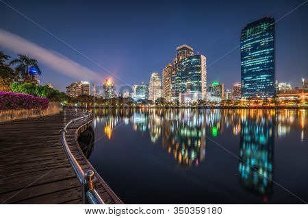 Skyscrapers And Lake With Wooden Walk Way In City Park. View Of Benjakiti Park At Night. Beautiful N