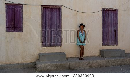 Asian Woman In Sunglasses And Hat Standing In Front Of Traditional Greek House With Purple Shutters 