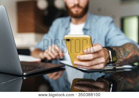 Close up of a concentrated young man using mobile phone while working on laptop computer in the kithcen at home