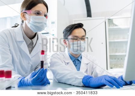 Two unrecognizable pharmacologists wearing protective lab coats, masks and gloves working together on new vaccine