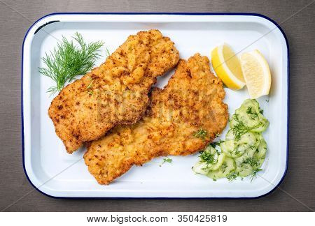 Traditional deep fried Wiener schnitzel from veal topside with cucumber salad and lemon slices offered as top view on a white tray 