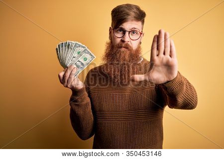 Redhead Irish man with beard holding a bunch of 1 dolla banknotes over yellow background with open hand doing stop sign with serious and confident expression, defense gesture
