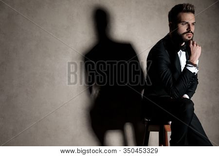 Thoughtful groom holding his hand on his chin and looking away while wearing tuxedo and sitting on a stool on wallpaper studio background