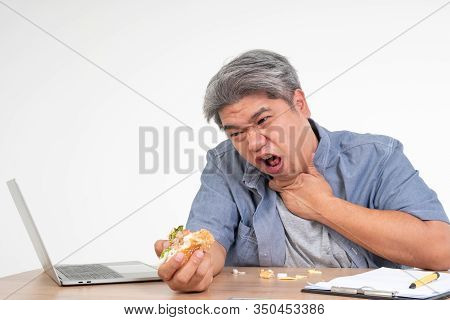 Asian Man Working And Eating A Burger On Office Desk And Holding His Neck After Choking Foods. Conce