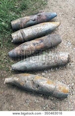 Five Unexploded Artillery Shells Lie On The Side Of The Asphalt Road, Dandelions Bloom And The Grass