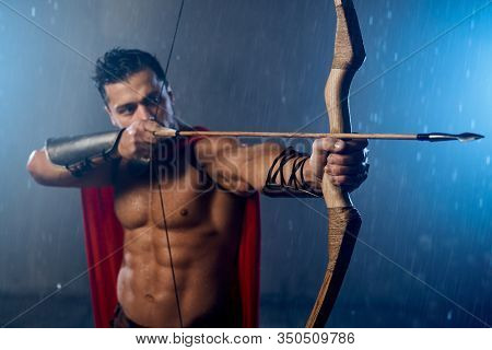 Front View Of Muscular Mature Spartan Wearing Red Cloak, Shooting From Bow With Arrows While Raining