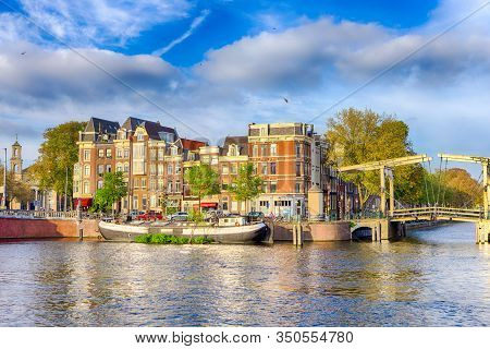 Traditional Dutch Cityscape Of Amsterdam With Barge In The Foreground At Daytime. Horizontal Image O
