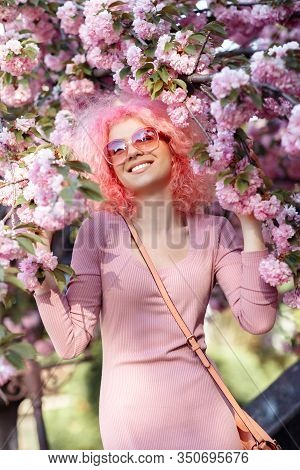Bright Woman With Curly Pink Hair And Sunglasses Near The Blossoming Spring Tree.