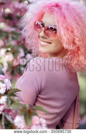 Close-up Portrait Of Pretty Young Woman With Curly Pink Hair And Sunglasses Near The Blossoming Spri