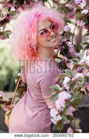 Close-up Portrait Of Bright Young Woman With Curly Pink Hair And Sunglasses Near The Blossoming Spri