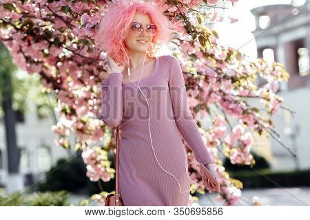 Attractive Woman With Curly Pink Hair And Sunglasses Near The Blossoming Spring Tree Listening Music