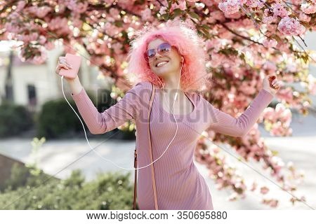 Happy Woman With Curly Pink Hair And Sunglasses Near The Blossoming Spring Tree Listening Music On S