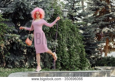 Dancing Young Woman With Curly Pink Hair And Sunglasses Near The Blossoming Spring Tree.