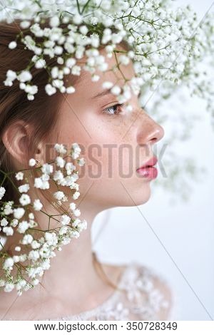 Beautiful tender girl with lovely freckles on her face and in a gypsophila wreath on a white background. Inspiration of spring and summer. Perfume, cosmetics concept. Prifile portrait. 
