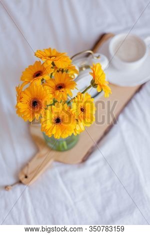 A Bouquet Of Gerberas In A Glass Jar, A Cup Of Cappuccino, A White Alarm Clock On A Wooden Stand Sta