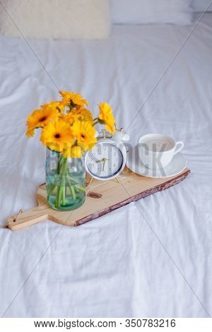 A Bouquet Of Gerberas In A Glass Jar, A Cup Of Cappuccino, A White Alarm Clock On A Wooden Stand Sta