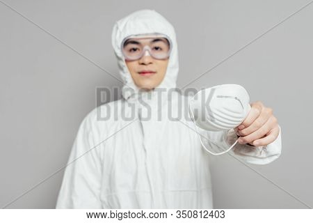 Asian Epidemiologist In Hazmat Suit Showing Respirator Mask While Looking At Camera On Grey Backgrou
