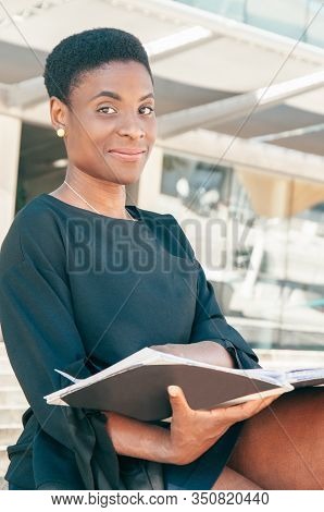 Pensive Smiling Black Woman Sitting On Stairs Outdoors And Turning Pages Of Album. Happy African Ame