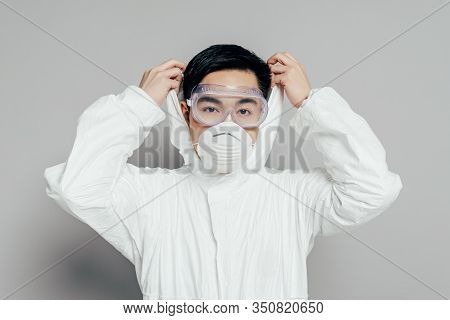 Asian Epidemiologist In Hazmat Suit And Respirator Mask Putting Hood On While Looking At Camera On G