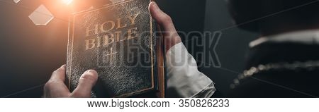 Partial View Of Catholic Priest Holding Holy Bible Near Confessional Grille In Dark With Rays Of Lig