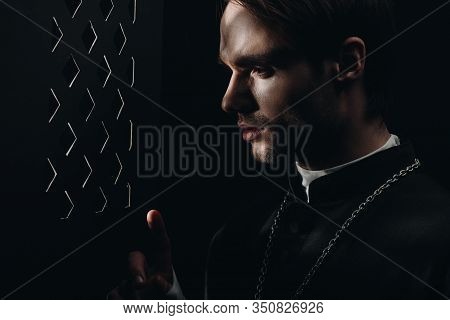 Young Tense Catholic Priest Looking Through Confessional Grille In Dark With Rays Of Light