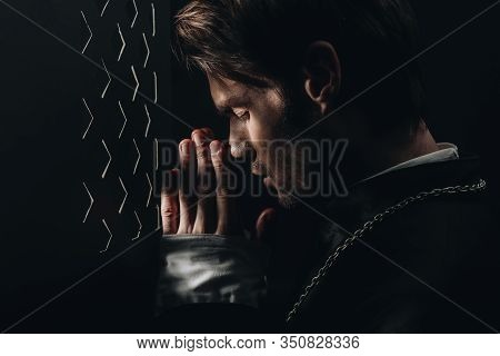 Young Catholic Priest Praying With Closed Eyes Near Confessional Grille In Dark With Rays Of Light