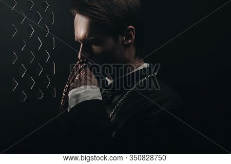 Young Thoughtful Catholic Priest Kissing Wooden Rosary Beads Near Confessional Grille In Dark With R