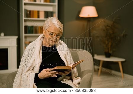 Woman Of Retirement Age Rests In Armchair Reading Old Book. Charming Gray-haired Lady Threw White Pl