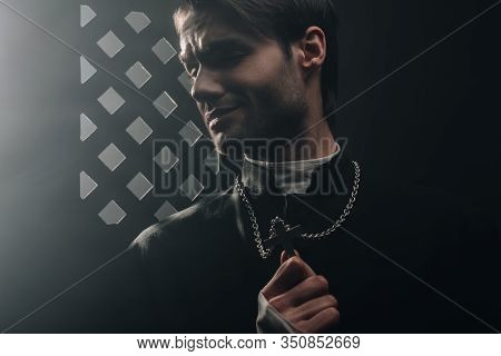 Young Worried Catholic Priest Touching Cross On His Necklace In Dark Near Confessional Grille