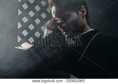 Young Thoughtful Catholic Priest Touching Face While Reading Bible Near Confessional Grille In Dark 