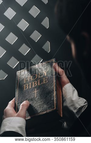 Partial View Of Catholic Priest Holding Holy Bible Near Confessional Grille In Dark With Rays Of Lig