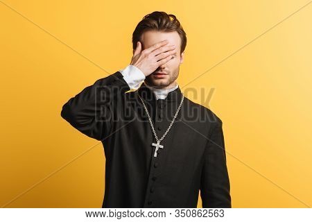 Young Catholic Priest Covering Eyes With Hand Isolated On Yellow