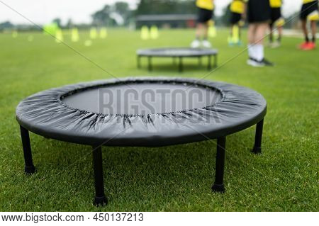 Fitness Training Trampoline On Grass Field. Football Team Standing In Blurred Background