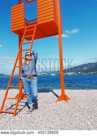 Happy Young Woman In Casual Clothes Standing On Beach On Sunny Summer Day Near Wooden Orange Lifegua