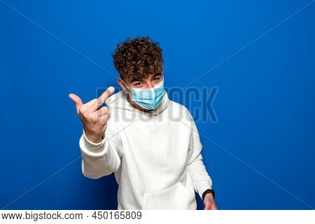 Portrait Of Angry Young Man In Surgical Medical Mask Showing Thumb Up And Looking At Camera With Ser