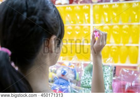 Asian Child Girl Holding A Dart And Aiming At The Balloons, Teen Playing The Dart Game For A Carniva