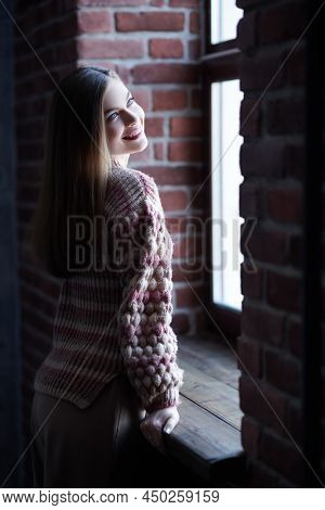 Portrait of an attractive happy girl in stylish casial clothes standing by a window in a loft room. Happy people and emotions. 