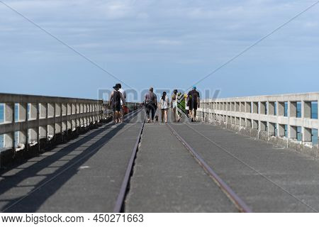 Tolaga Bay New Zealand - February 5 2022; Group People Walking Along Long Wharf With One Carrying Su