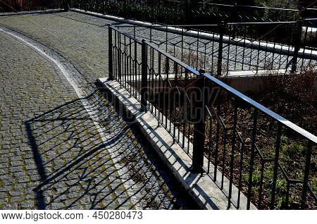 Paved Sidewalk With Gutter And Gray Metal Ornate Railings. Lamp With Glazed Lampshade And Black Post