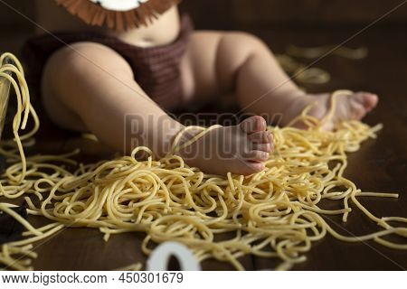 Little Chubby Kids Foot In Pasta On A Wooden Brown Background, Table. Feeding Concept. Spaghetti Sma